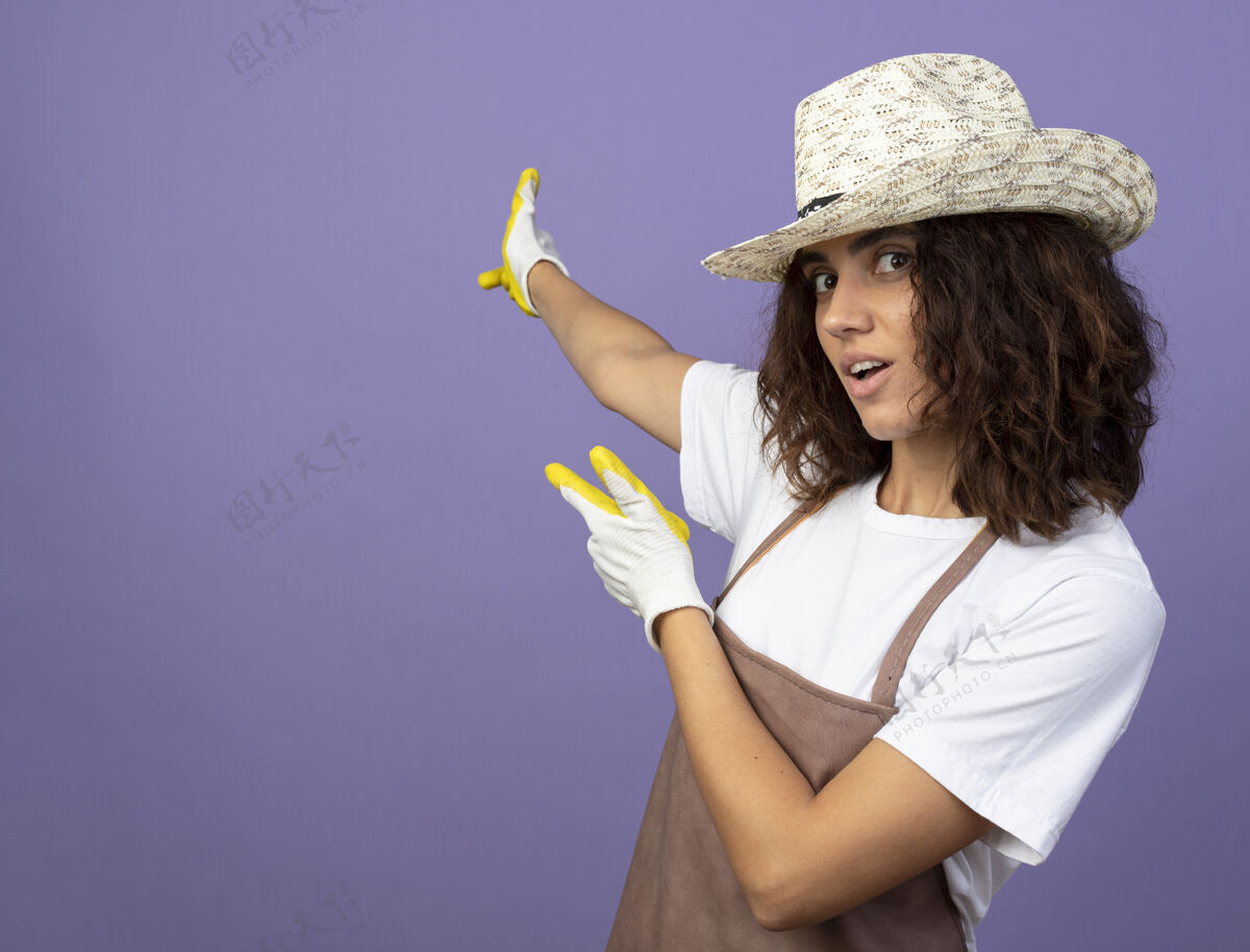 紫色高兴的年轻女园丁穿着制服 戴着园艺帽和手套点在后面手套请制服