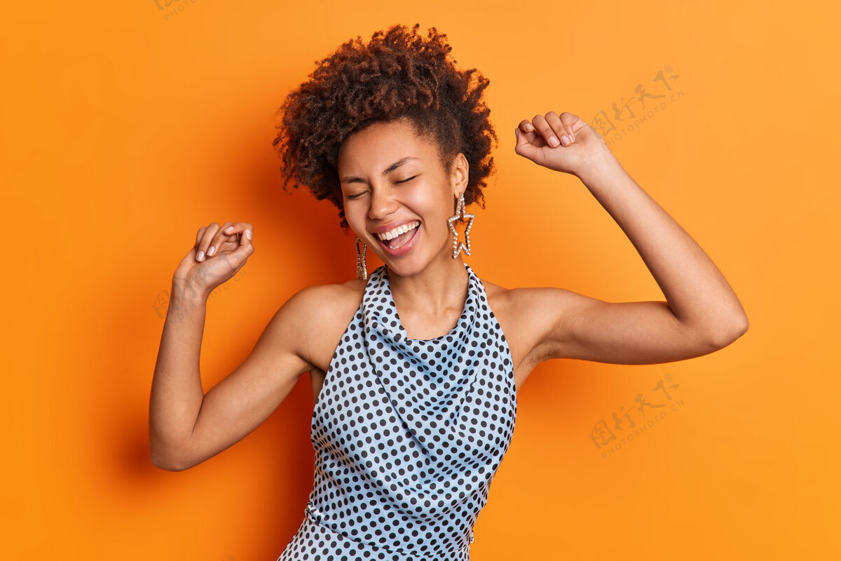 女孩摄影棚拍摄的无忧无虑的年轻美籍黑人女子动作积极地举起双臂 感觉充满活力的笑容广泛地在迪斯科派对上度过空闲时间 在橙色的摄影棚背景下与世隔绝大笑娱乐积极