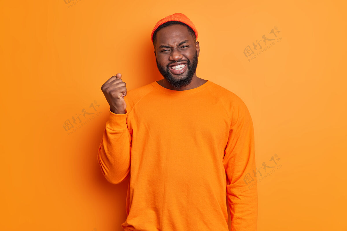 力量情绪化的黑皮肤胡须男人咬紧牙关 举起拳头表达对橙色背景上孤立的事物不满的负面情绪强硬年轻攻击