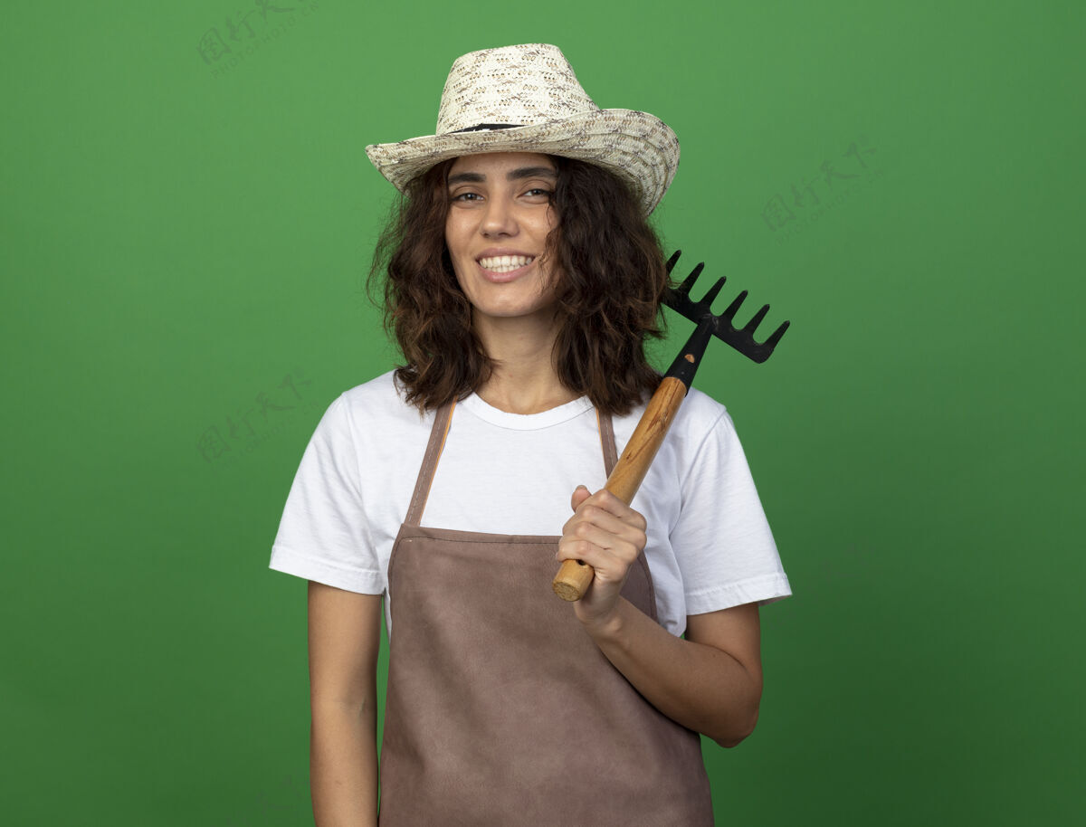 女人身着制服的年轻女园丁微笑着戴着园艺帽 肩上扛着耙子年轻人微笑穿
