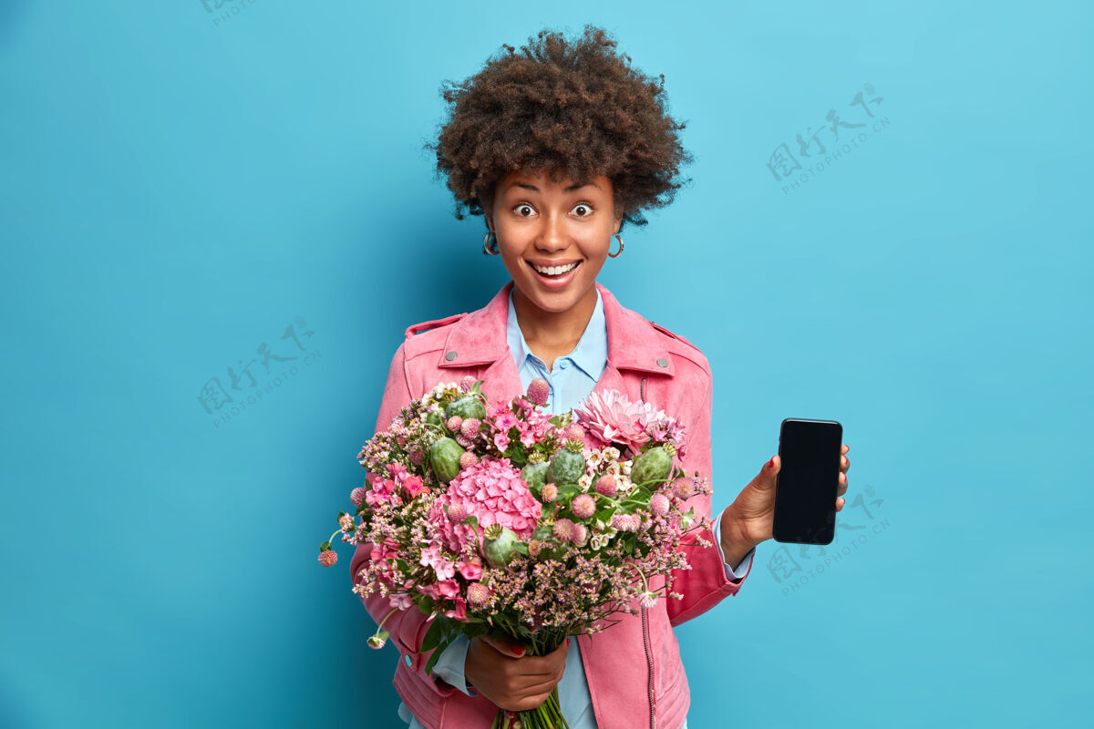 花开朗的非洲裔美国年轻女子手持花束展示智能手机与实物模型展示微笑积极享受特殊的节日欢呼肖像牙关