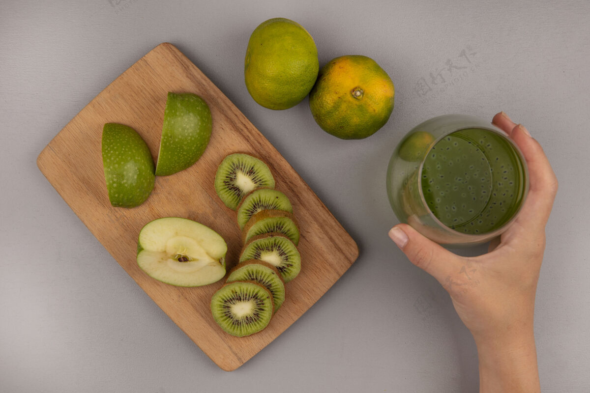 水果顶视图中的女性手拿着一杯新鲜的猕猴桃汁和切碎的苹果片猕猴桃放在木制的厨房板上与橘子隔离雌性视图板
