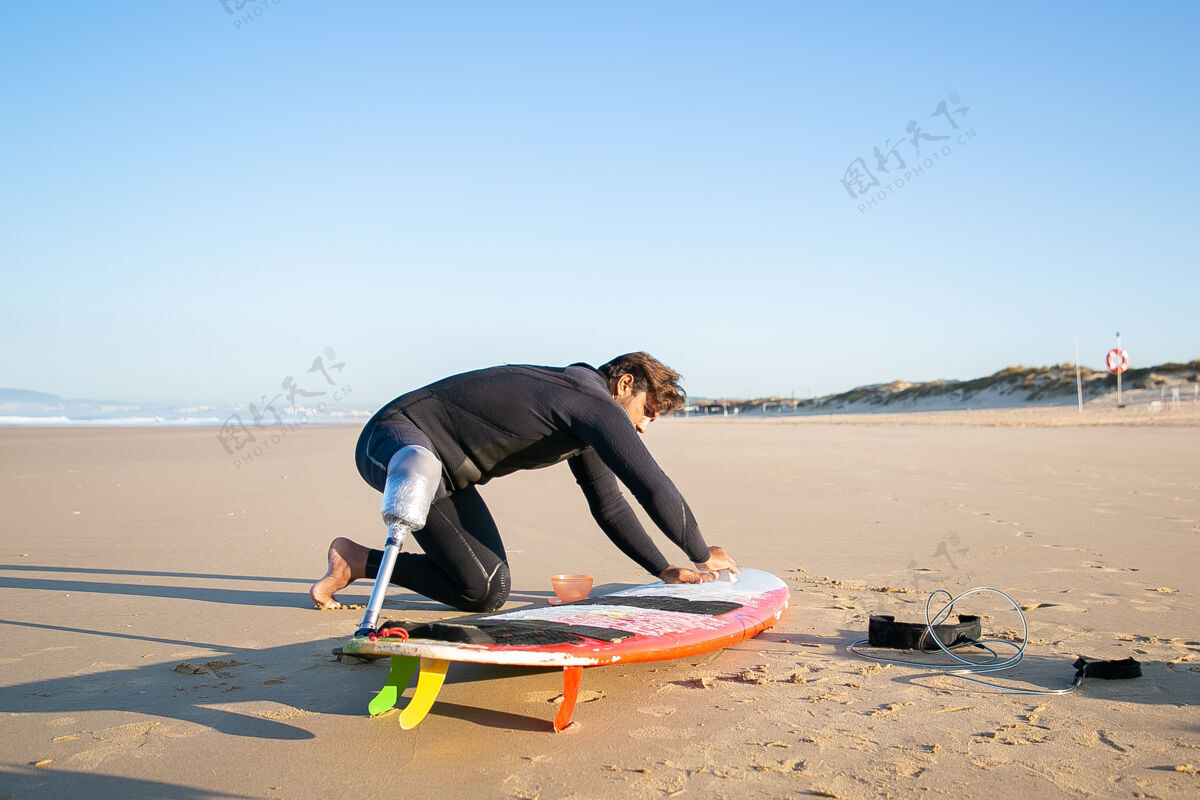 肖像穿着潜水服的冲浪者戴着假肢 在沙滩上给冲浪板打蜡肢体残疾冲浪者