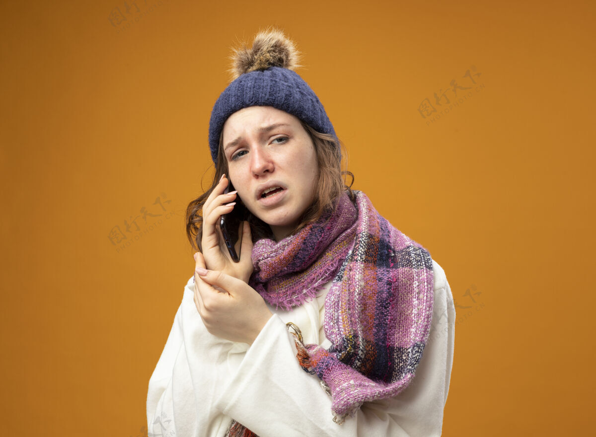 年轻穿着白色长袍 戴着带围巾的冬帽 在橘色墙上孤零零地讲着电话的未出柜的生病的年轻女孩冬天女孩说话