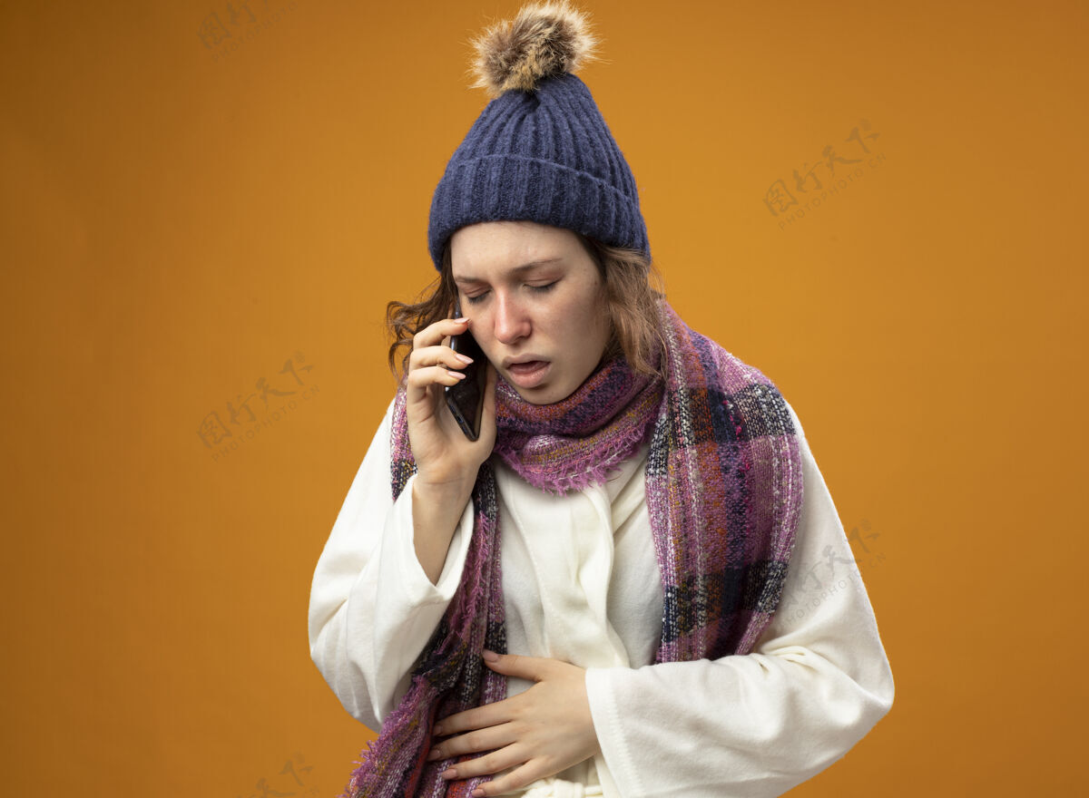 手咳嗽的年轻病女孩穿着白色长袍 戴着带围巾的冬帽 手放在肚子上讲电话 隔离在橙色的墙上疾病电话围巾