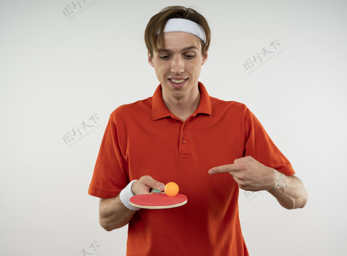 点高兴的年轻人戴着带腕带的头带 指着乒乓球拍 球被隔离在白墙上乒乓球乒乓球年轻