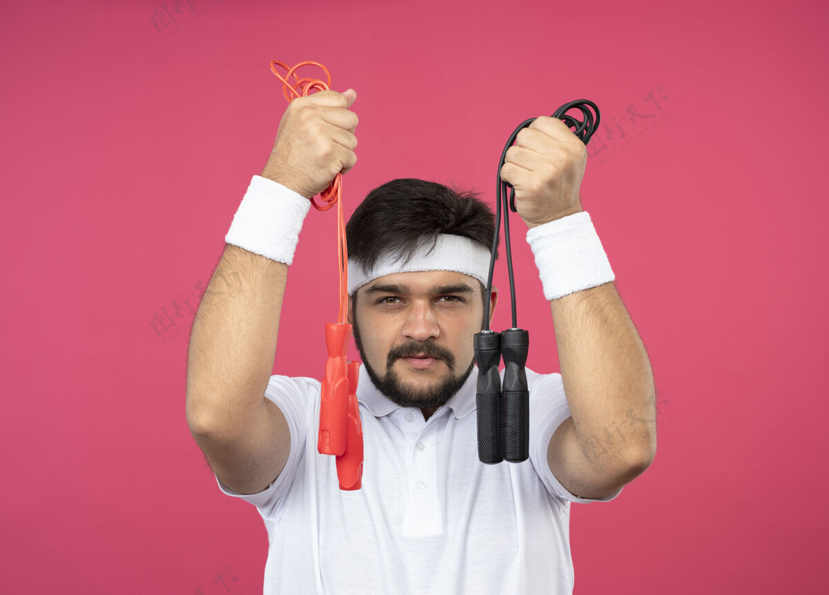 手持高兴的年轻运动男子戴着头带和手环举行跳绳隔离粉红墙上绳索腕带年轻