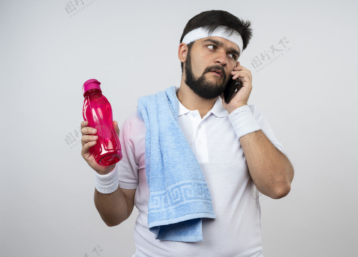 瓶子困惑的年轻人戴着头带和腕带肩上拿着毛巾拿着水瓶在电话里讲话隔离在白墙上运动穿肩膀