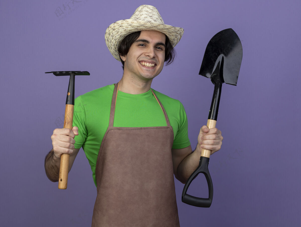 持有微笑着的年轻男园丁身着制服戴着园艺帽拿着耙子用铁锹孤立在紫色地上紫色耙制服