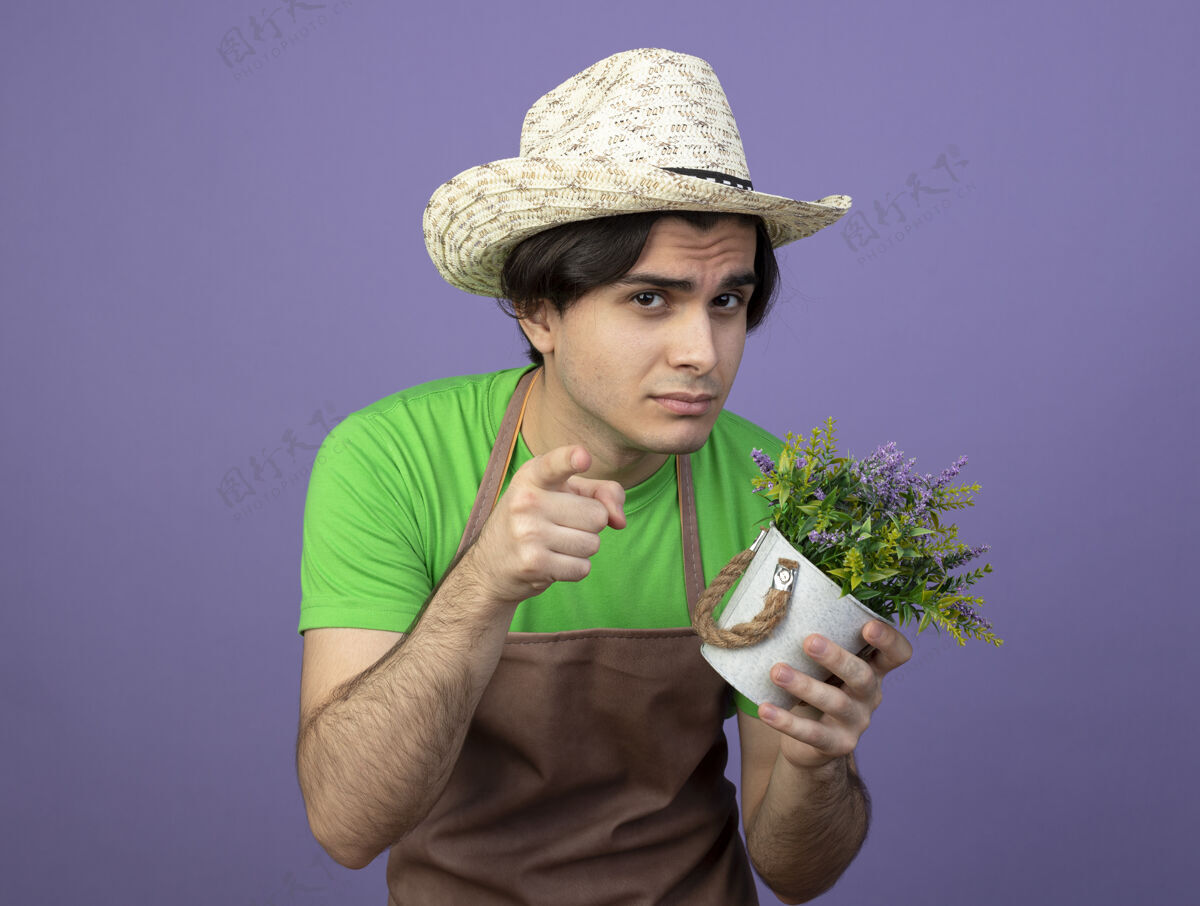 举行可疑的年轻男园丁穿着制服 戴着园艺帽 手里拿着花盆里的花 向你展示紫色的孤立姿态男性手势制服