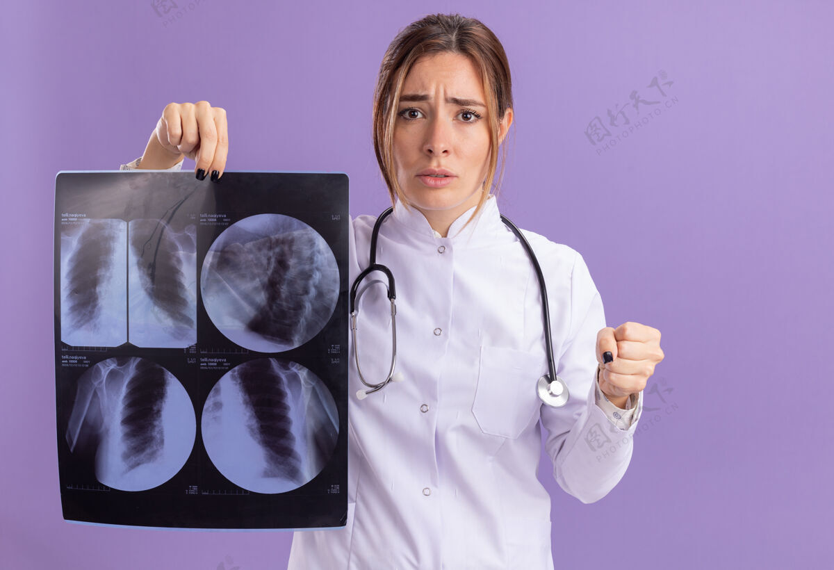 医疗年轻严格的女医生 穿着医用长袍 手持听诊器 手持x光片 手持拳头 隔离在紫色的墙上严格听诊器年轻