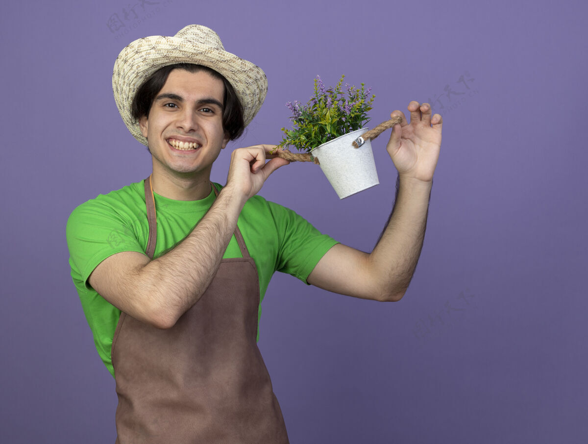 小伙子微笑着的年轻男园丁穿着制服 戴着园艺帽 手里拿着紫罗兰上孤立的花盆里的花加油