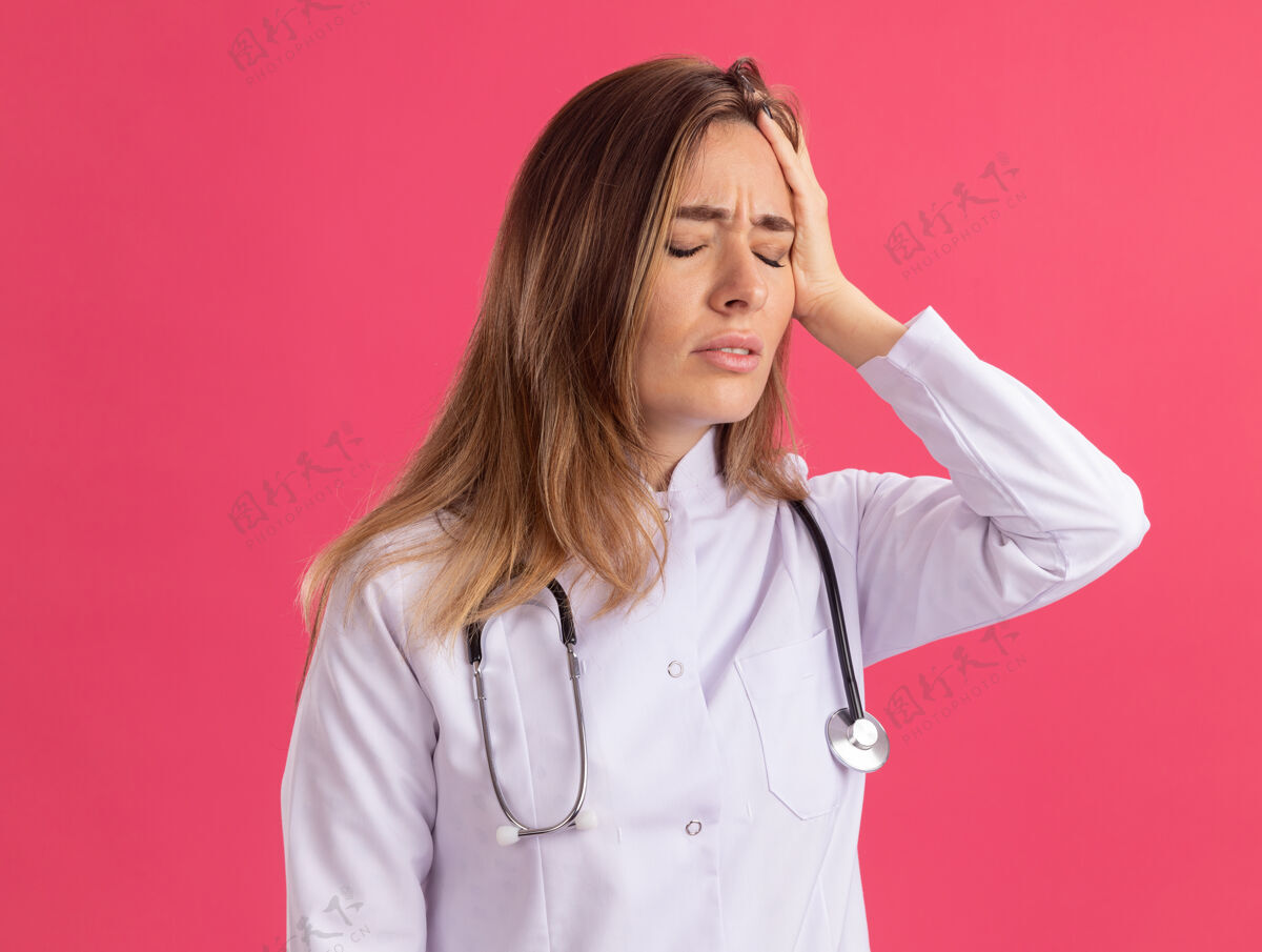 封闭闭着眼睛疼痛年轻女医生穿着医用长袍听诊器把手放在头上隔离在粉红色的墙上听诊器穿着手