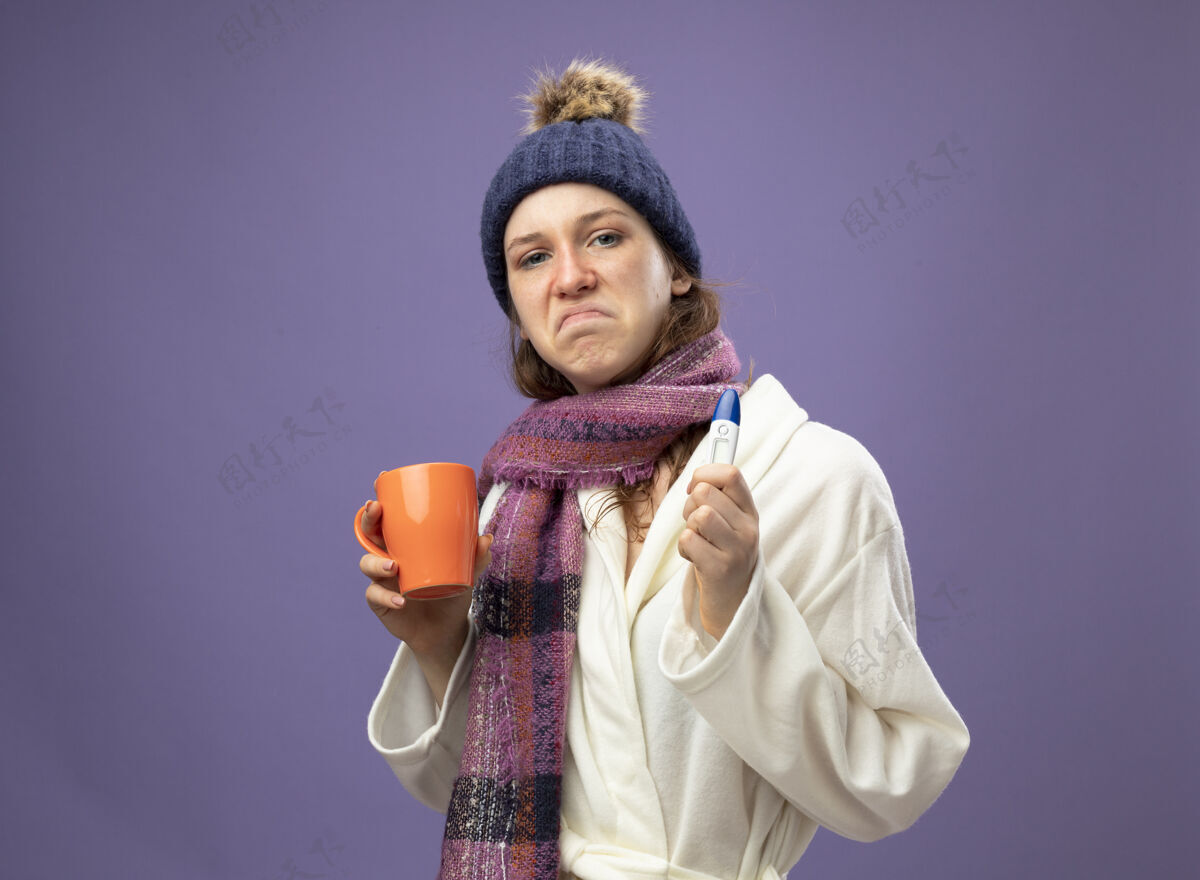 帽子悲伤的生病的年轻女孩穿着白色长袍 戴着冬天的帽子 戴着围巾 手里拿着一杯茶 温度计隔离在紫色上体温计茶杯子