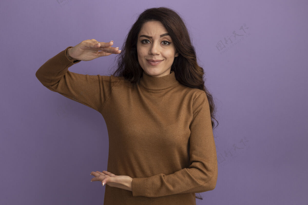 毛衣可爱的年轻漂亮女孩穿着棕色高领毛衣 在紫色的墙上显示出孤立的尺寸展示高领毛衣穿