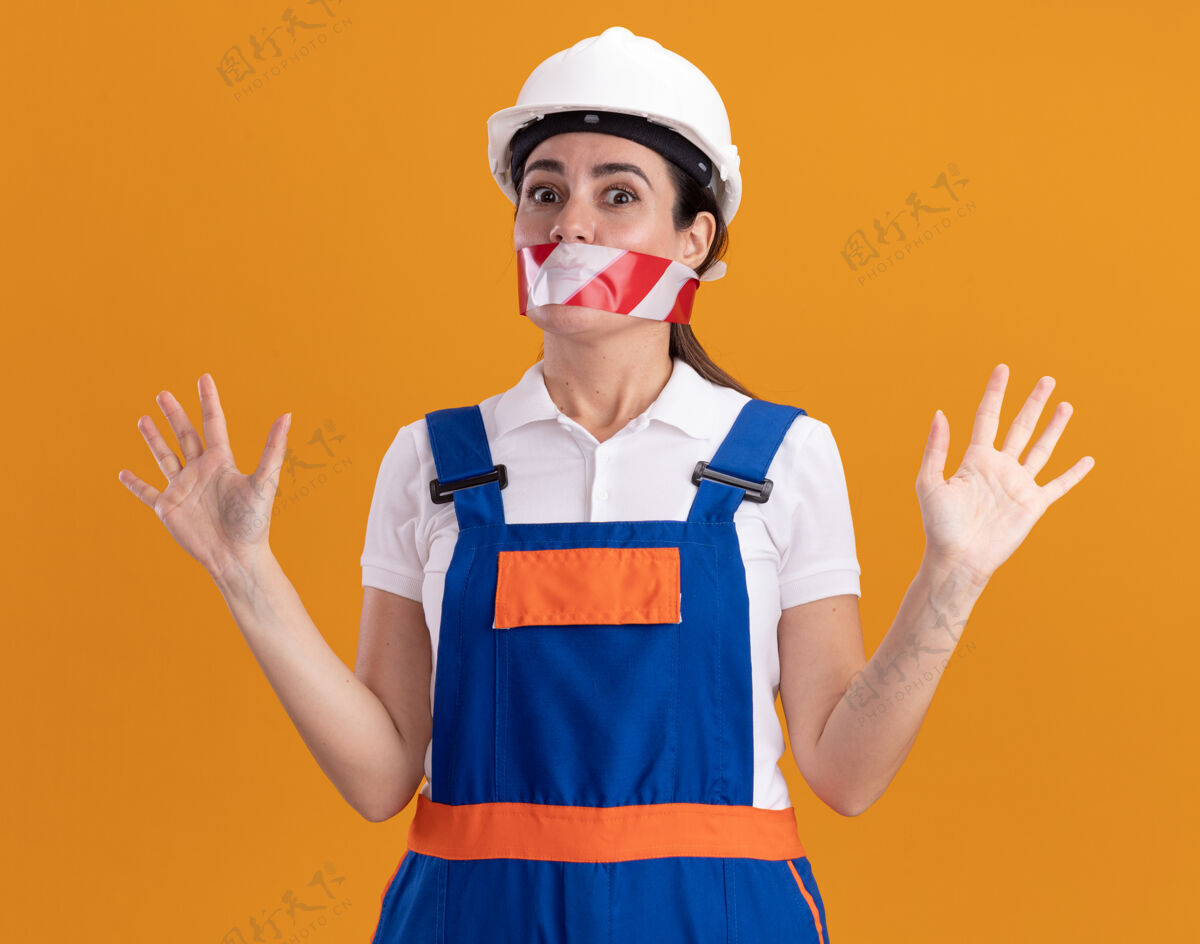 胶带穿着制服的年轻建筑工人惊讶地闭上了嘴 用胶带把双手分开放在橙色的墙上传播管道建筑工人