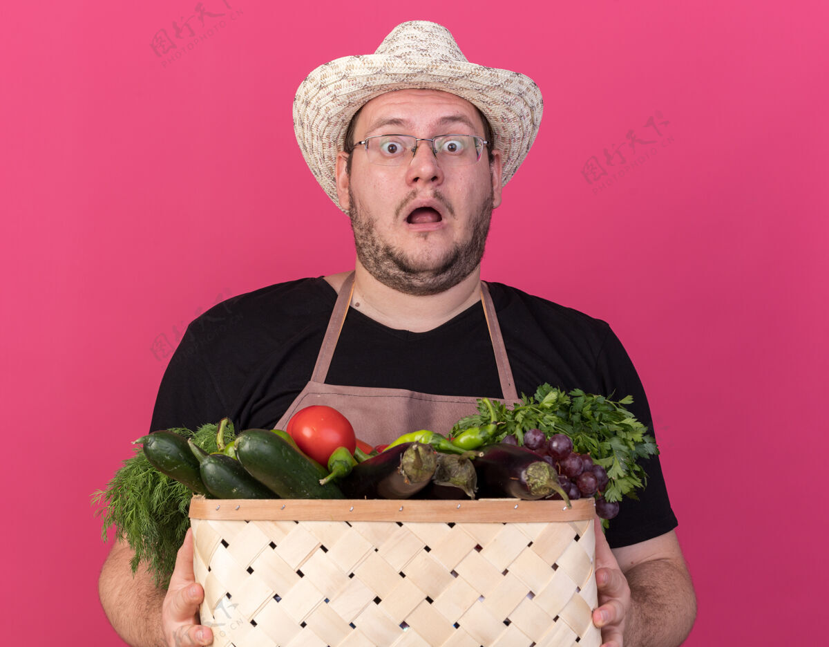 花园被吓坏了的年轻男园丁戴着园艺帽拿着蔬菜篮子孤立在粉红色的墙上害怕园艺穿