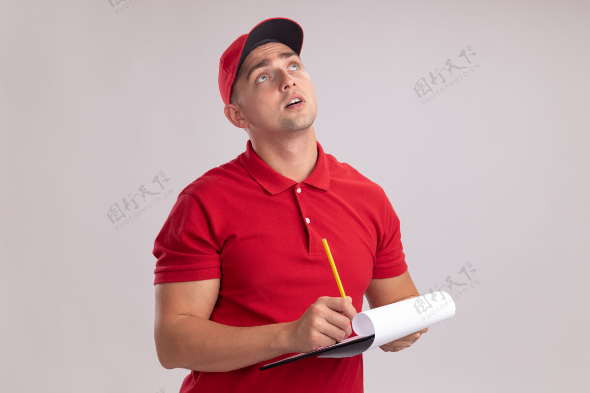 铅笔令人印象深刻的年轻送货员穿着制服 戴着帽子 拿着铅笔的剪贴板 隔离在白色的墙上戴着剪贴板帽子