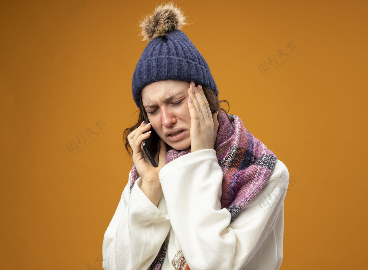 太阳穴哭泣的生病的年轻女孩穿着白色长袍 戴着冬天的帽子 戴着围巾 讲着电话 把手放在橘子色的太阳穴上穿疾病电话