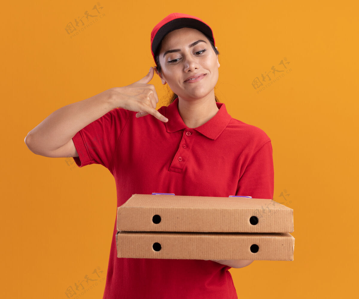 电话穿着制服 戴着帽子 拿着披萨盒 在橙色的墙上显示着电话的手势 年轻的送货女孩很高兴电话盒子帽子