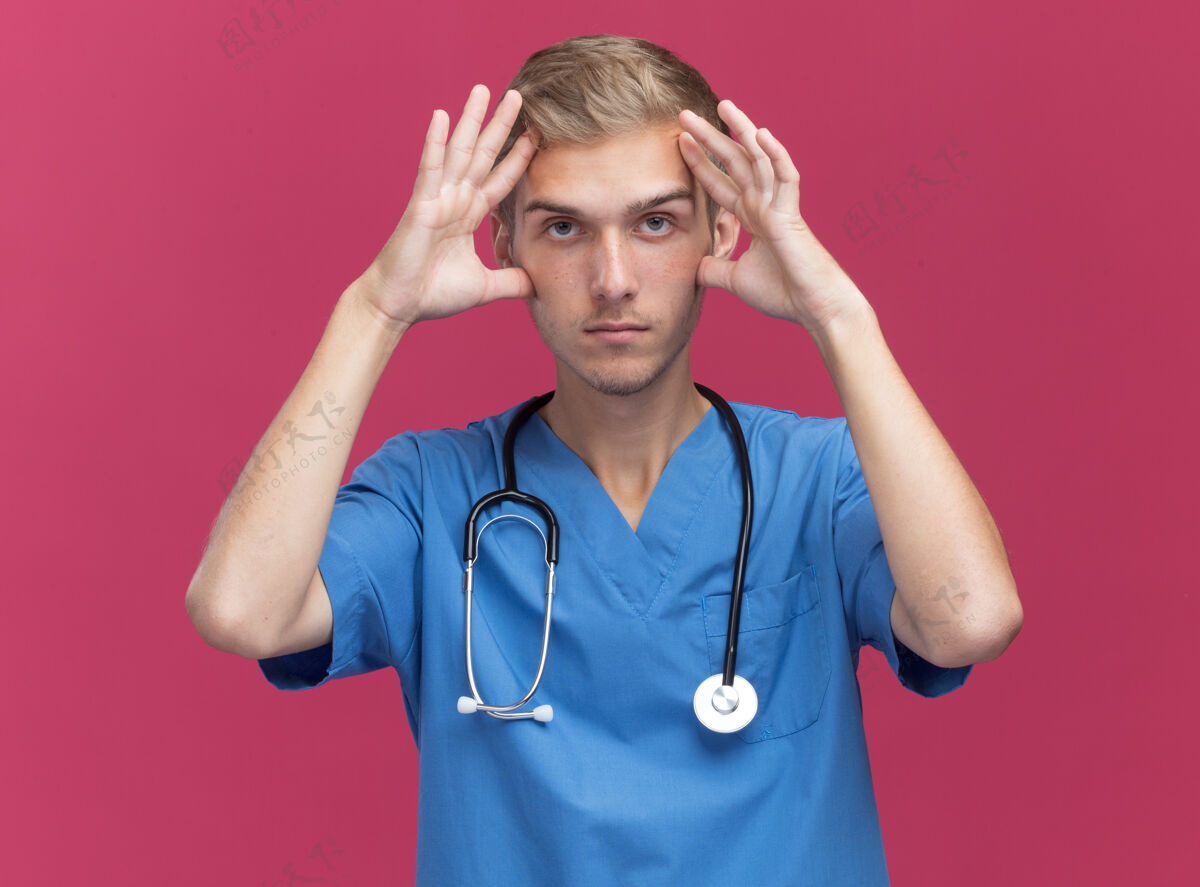手疲惫不堪的年轻男医生穿着医生制服 手放在隔离在粉红色墙上的前额上 戴着听诊器医生疲倦戴