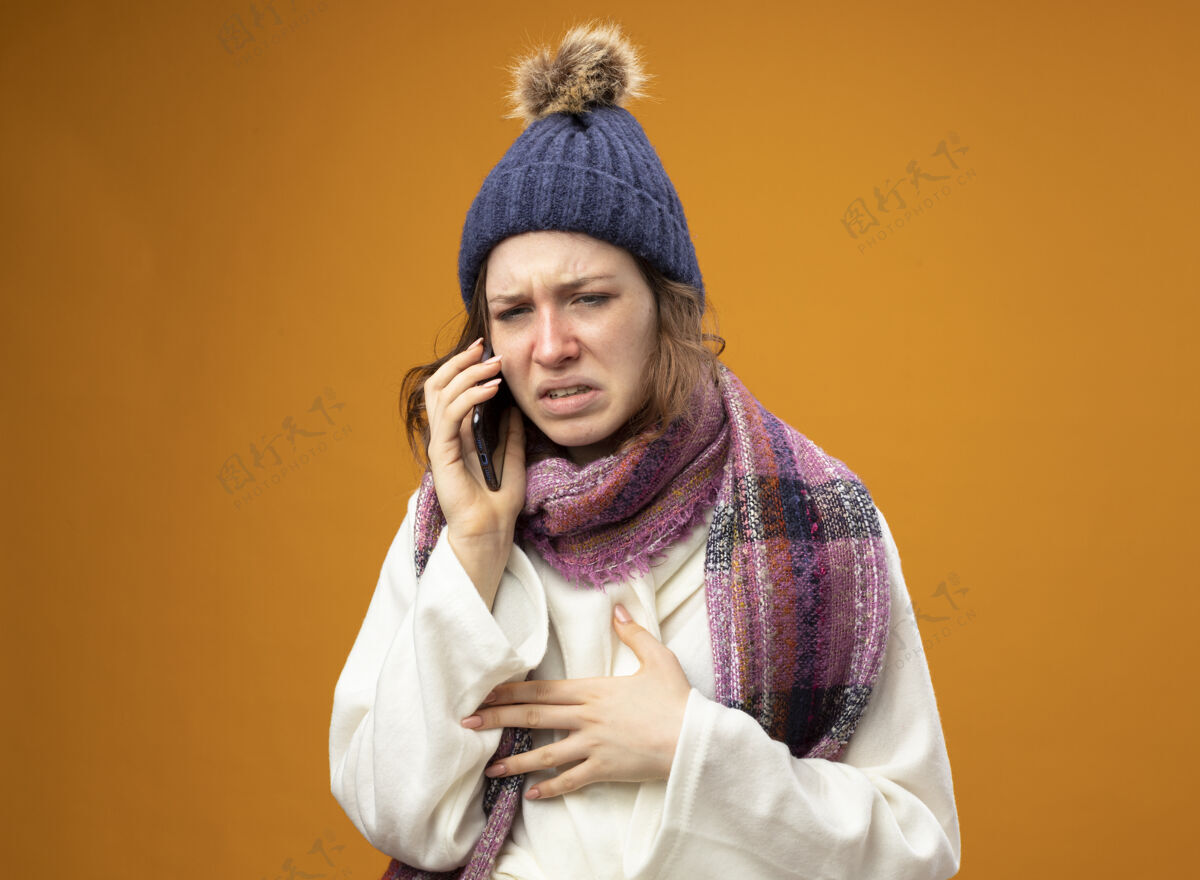 橙色穿着白色长袍 戴着带围巾的冬帽的虚弱的生病的年轻女孩在电话里说橙色的孤立长袍穿疾病
