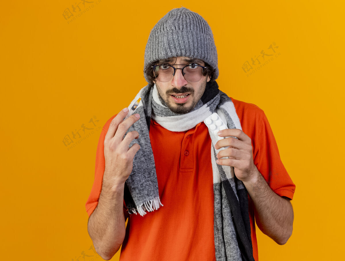 穿着印象深刻的年轻病人戴着眼镜 戴着冬天的帽子 戴着围巾 拿着注射器和一包药片 看着前面隔离着的橘黄色墙壁 留着复印空间围巾医学感觉