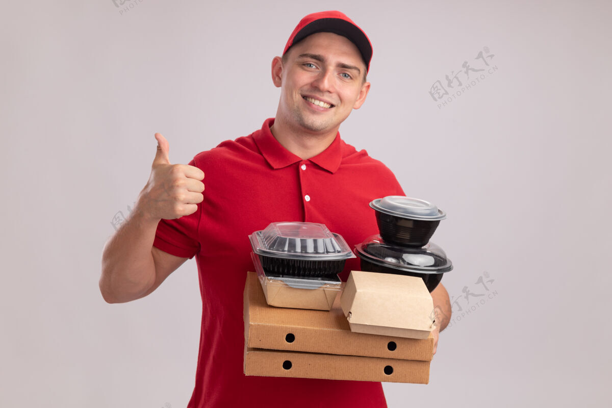 拇指微笑的年轻送货员身穿制服 戴着帽子 拿着比萨饼盒上的食品容器 在白色的墙上孤立地竖起大拇指帽子盒子送货