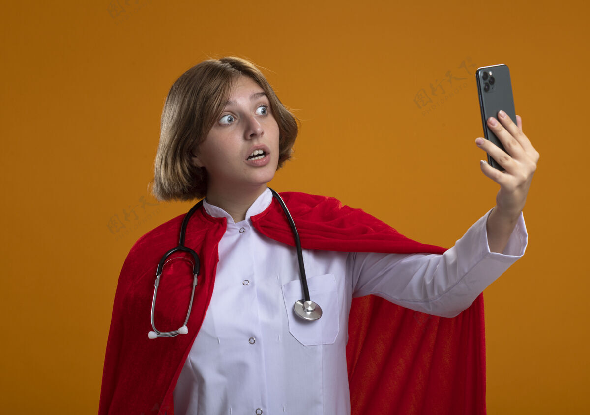 印象穿着医生制服和听诊器的穿着红色斗篷的金发女超人在橙色的墙上自拍金发感情市民