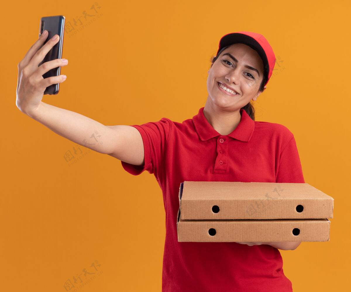 送货微笑着的年轻送货女孩穿着制服 戴着帽子 拿着披萨盒 在橙色的墙上独自自拍拿着披萨帽子