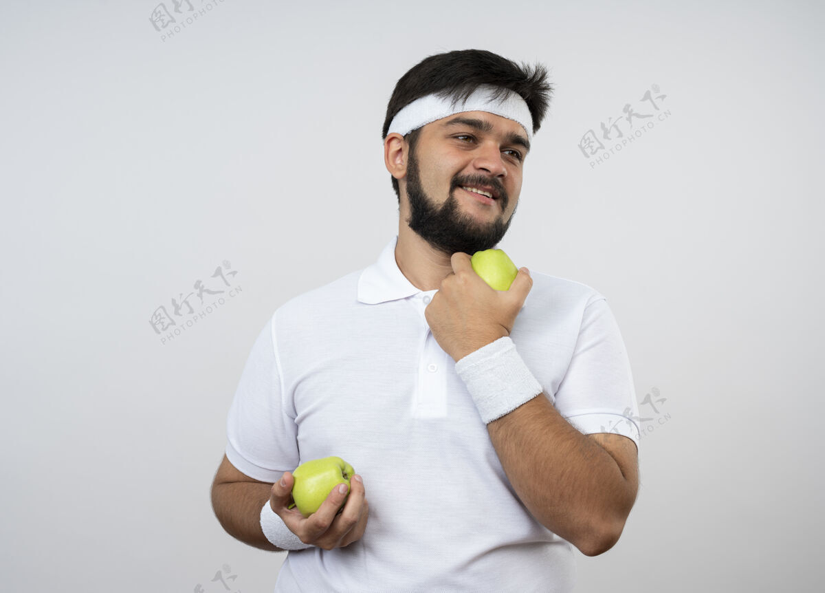 头带面带微笑的年轻运动型男子看着戴着头带和手环的一边 手里拿着孤立在白墙上的苹果苹果穿抱着
