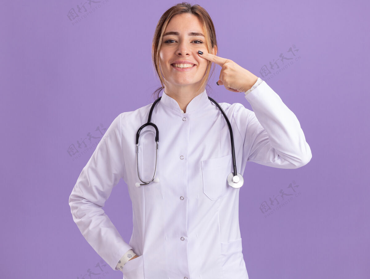 医生微笑的年轻女医生穿着医用长袍 用听诊器指着隔离在紫色墙上的眼睛听诊器长袍点