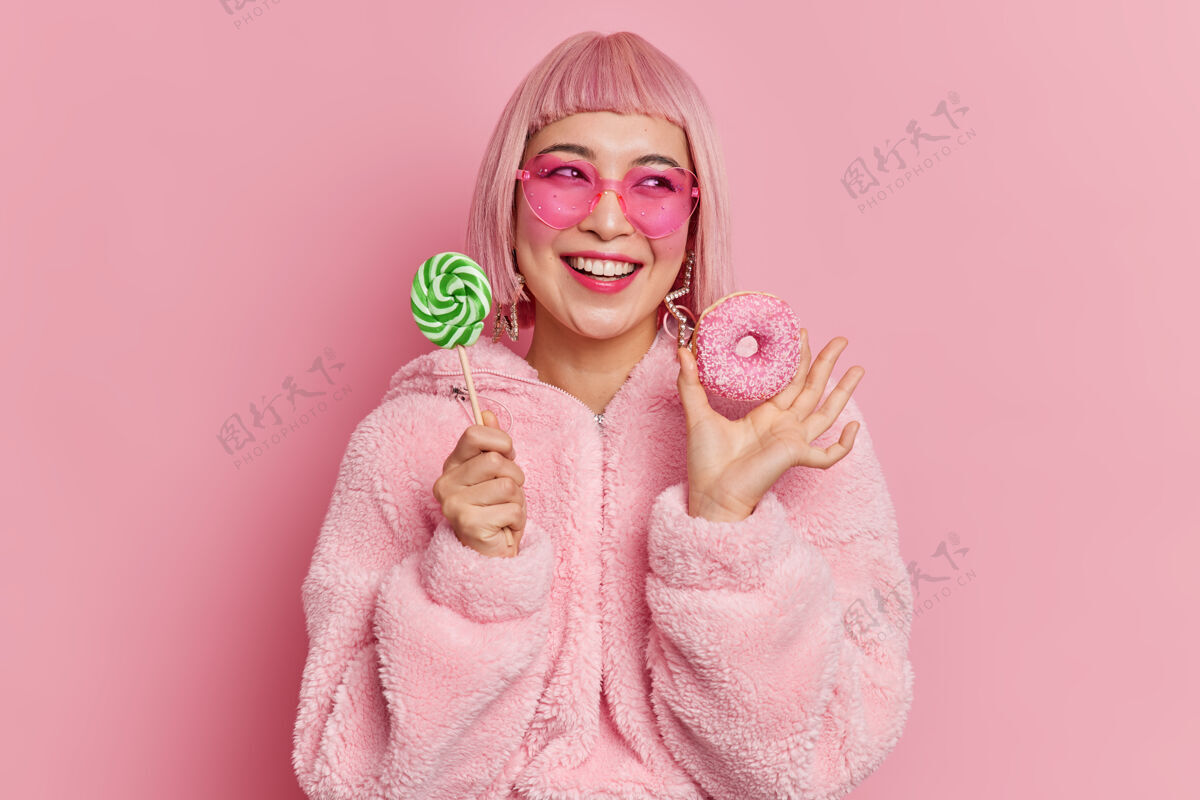 粉色笑容可掬的亚洲少女兴高采烈地把棒棒糖放在棒棒糖和开胃甜甜圈旁边时尚思考请