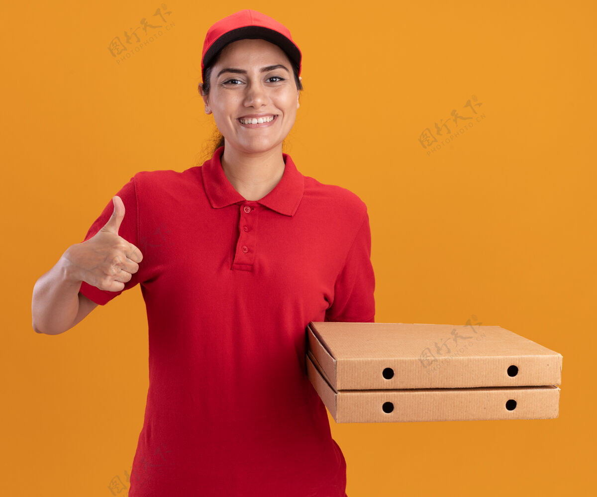 展示微笑的年轻送货女孩穿着制服 戴着帽子 拿着披萨盒 在橙色的墙上孤立地竖起大拇指穿盒子披萨