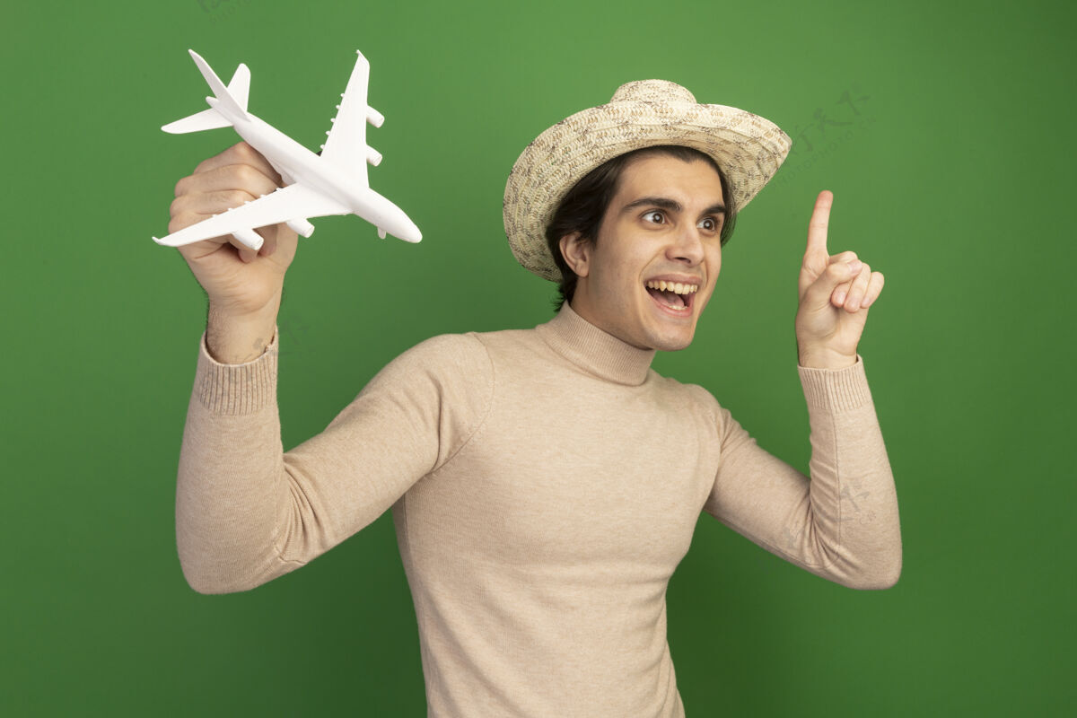 提高印象深刻的看着身边戴着帽子的年轻帅哥 举起玩具飞机指着绿色的墙上孤立起来穿点年轻