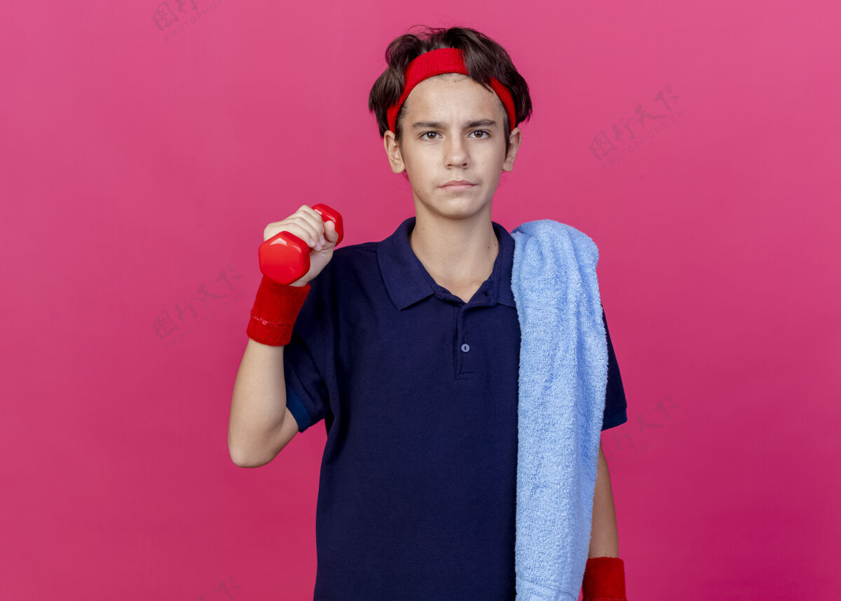 站着年轻帅气的运动男孩戴着头带和护腕 肩上戴着牙套和毛巾 看着前面拿着哑铃 隔离在粉红色的墙上 留着复制空间抱着人脸