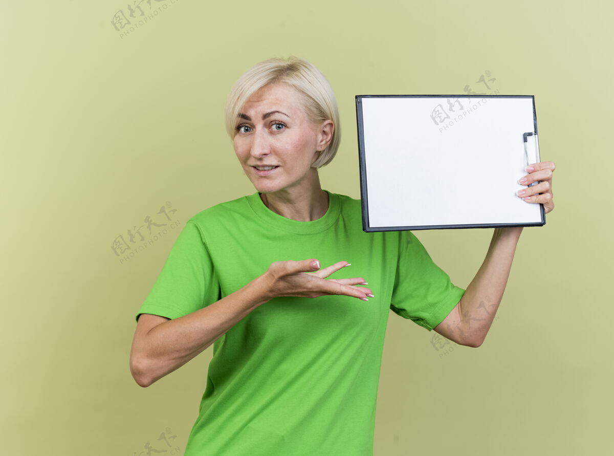 脸印象深刻的中年金发斯拉夫妇女拿着剪贴板靠近头部 用手指着它 看着前面孤立的橄榄绿墙上衣服靠近拿着