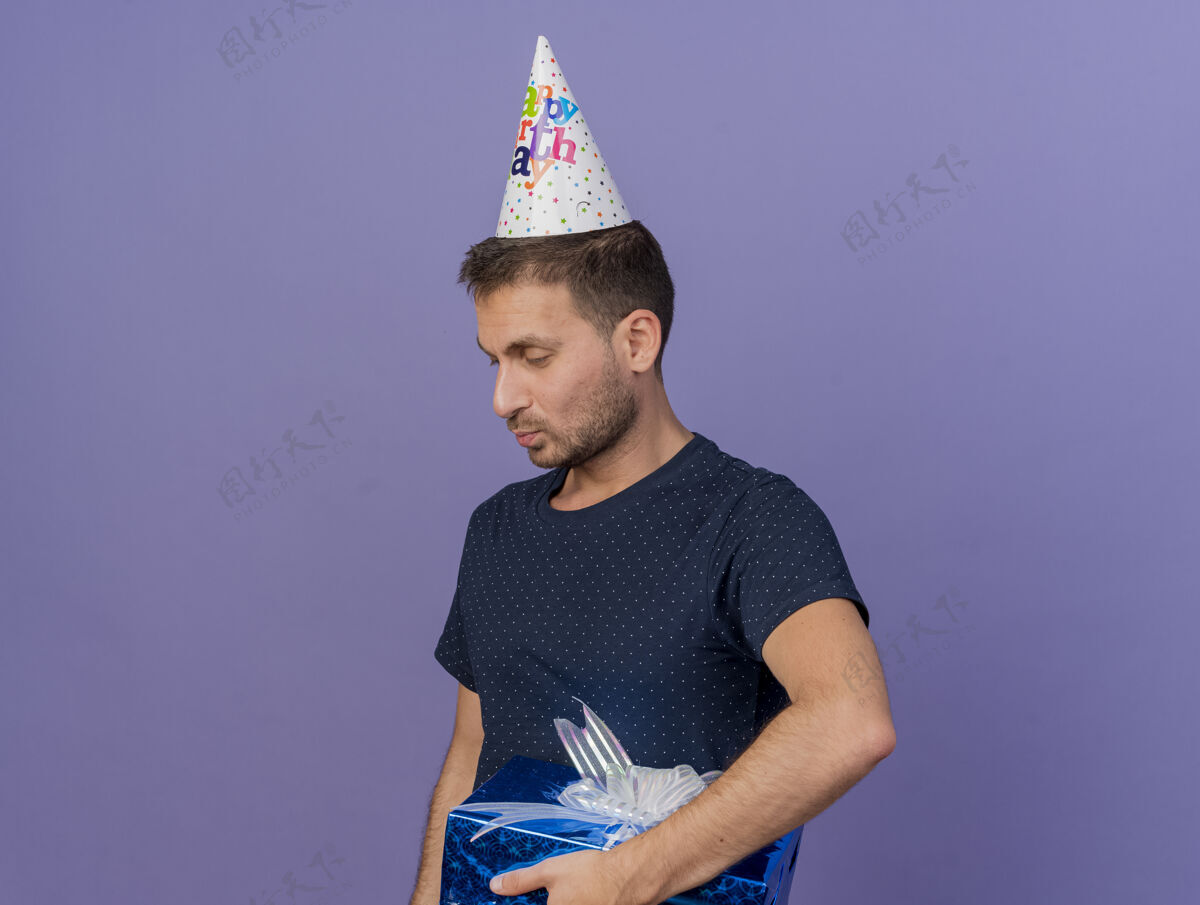盒子高兴的帅哥戴着生日帽拿着礼盒望着一面隔离在紫色墙上的复制空间感情表情请