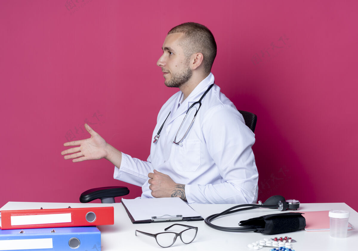 长袍年轻的男医生穿着医用长袍和听诊器坐在办公桌旁 带着工作工具转向一边看 伸出手在粉色背景上孤立地打招呼听诊器年轻你好