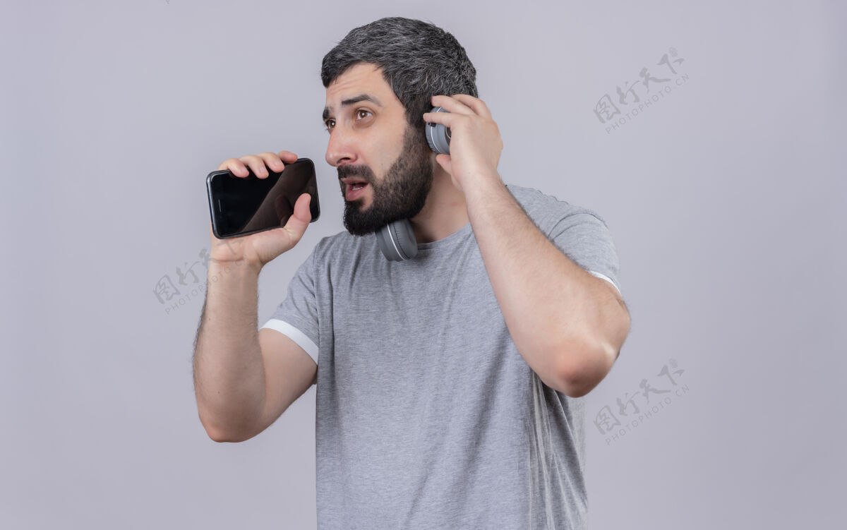 手机年轻帅气的白人男子戴着耳机假装唱歌 用手机当麦克风 一边看着一边用手戴着耳机 隔离在白色背景上 留有复印空间年轻耳机穿
