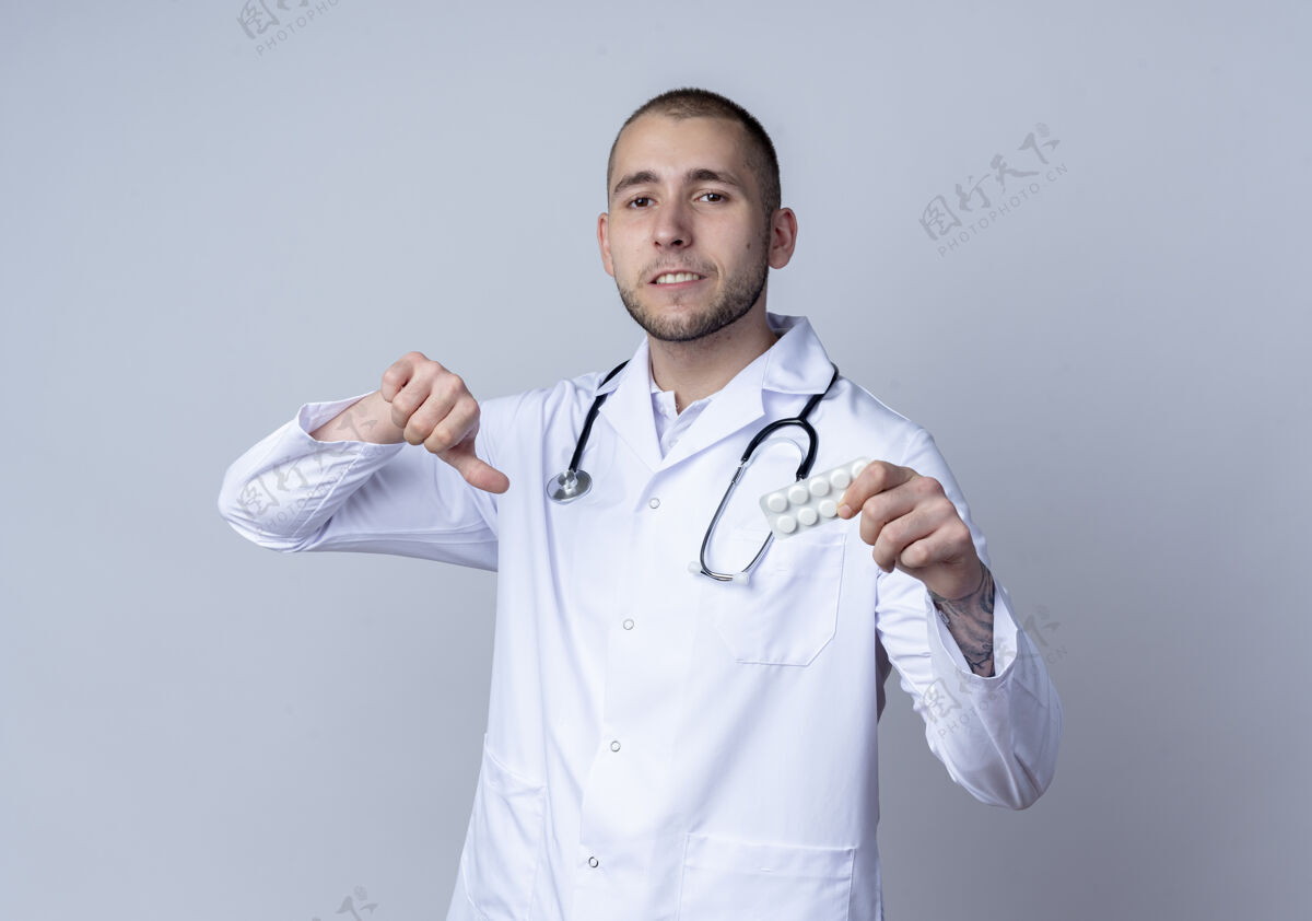 包年轻的男医生 穿着医用长袍 脖子上戴着听诊器 手里拿着一包药片 大拇指朝下 被隔离在白色背景上听诊器按住不愉快