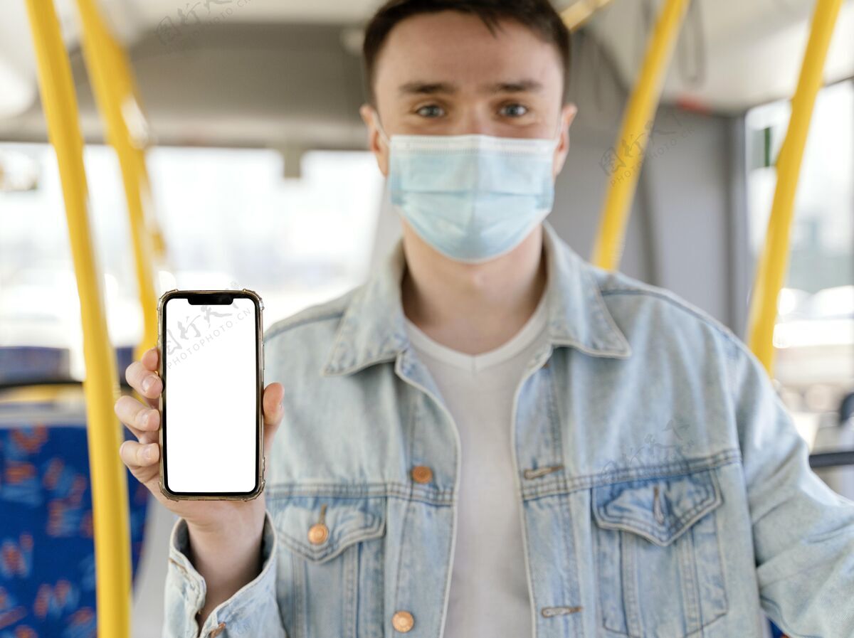 防护口罩坐城市巴士的年轻人在展示白屏智能手机手机交通旅行
