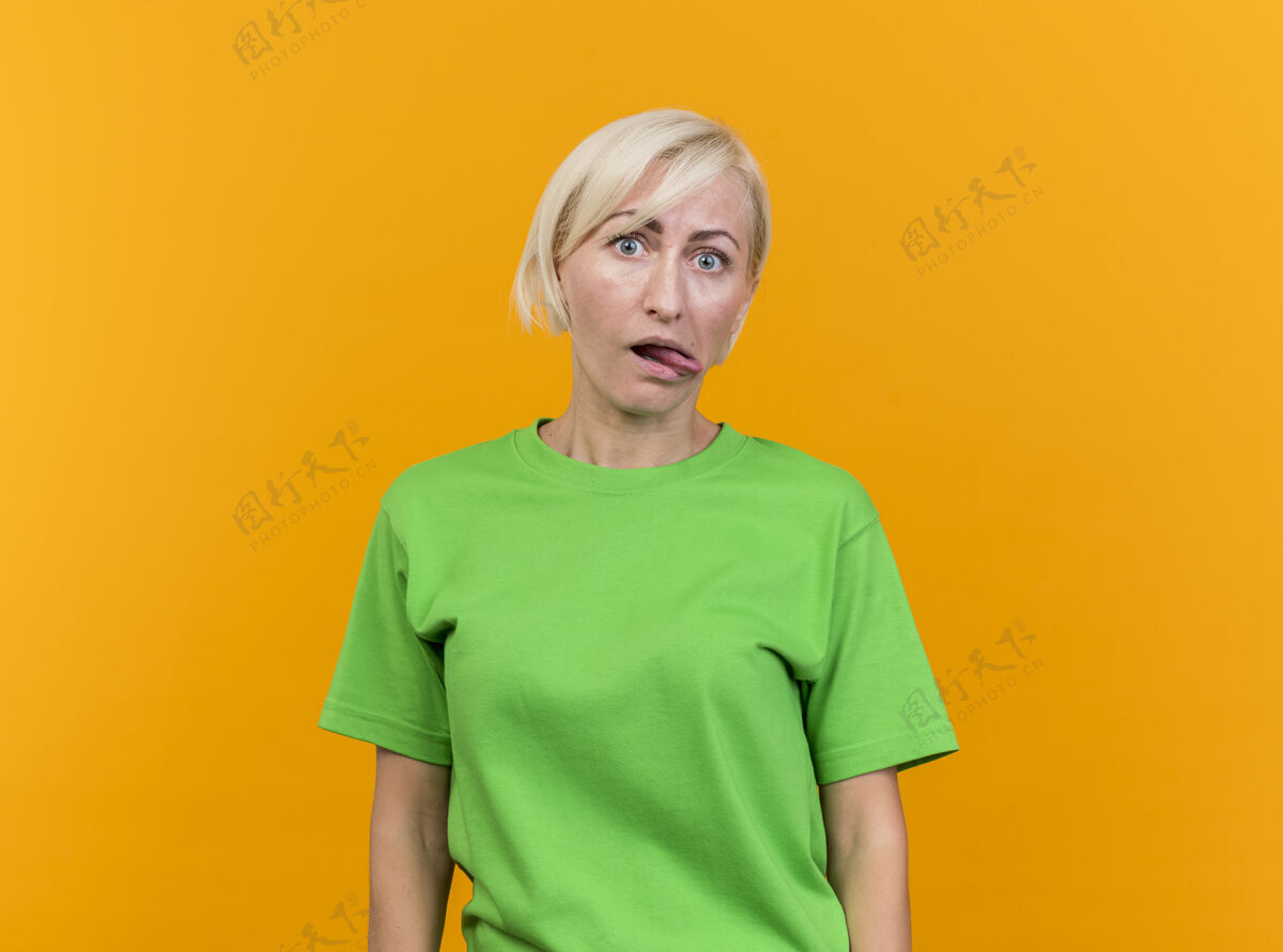 感觉顽皮的中年金发斯拉夫女人在看前面显示舌头隔离在黄色墙上与复制空间衣服人舌头