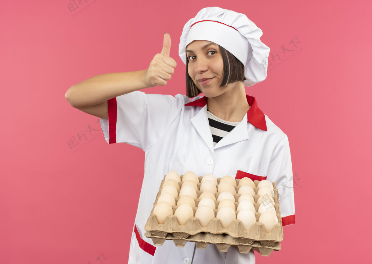 拿着身着厨师制服的年轻女厨师手捧一盒鸡蛋 在粉色背景上孤立地竖起大拇指制服厨师女
