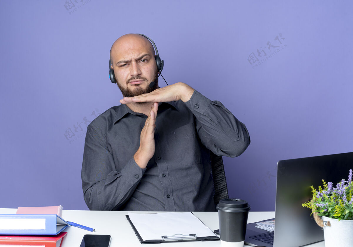 手势戴着耳机的秃头呼叫中心年轻人坐在办公桌旁 拿着工作工具做着超时手势 紫色背景下被隔离通话工作磨损