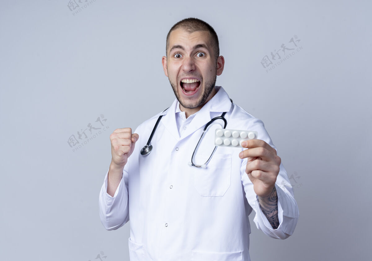 听诊器令人印象深刻的年轻男医生穿着医用长袍 脖子上戴着听诊器 伸出一包药片 紧握着隔离在白色皮肤上的拳头颈部穿着医生