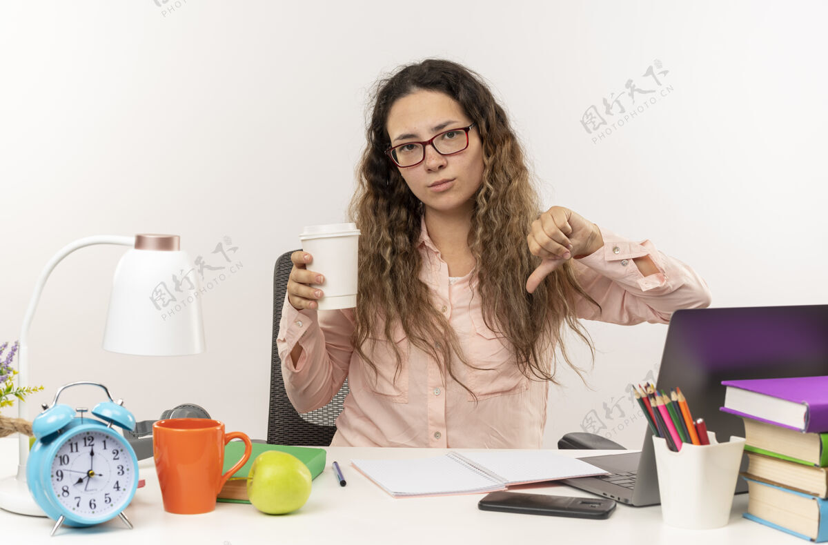 学校年轻漂亮的女学生戴着眼镜坐在课桌旁 手里拿着学习用具做作业 手里拿着咖啡杯 在白色背景上孤立地竖起大拇指不愉快漂亮拇指