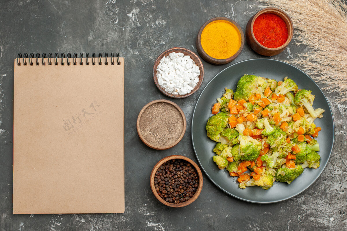 晚餐健康餐 黑盘子上放花椰菜和胡萝卜 灰色桌上笔记本旁放香料蔬菜胡椒午餐