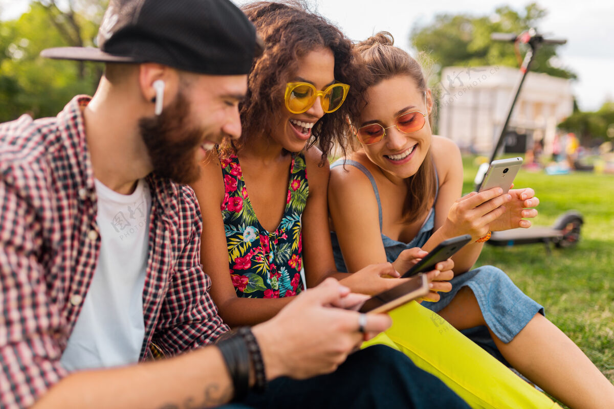 多种族快乐的年轻朋友们坐在公园里用智能手机微笑 男人和女人一起玩享受音乐女人