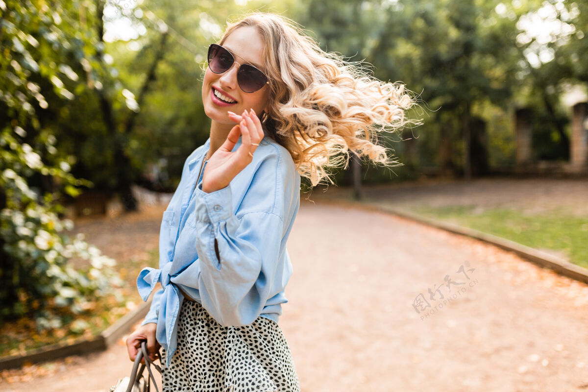 时尚迷人的金发微笑的女人在公园散步 夏天穿蓝色衬衫和裙子 戴着优雅的太阳镜和钱包 街头时尚风格 心情愉快享受手提包太阳镜